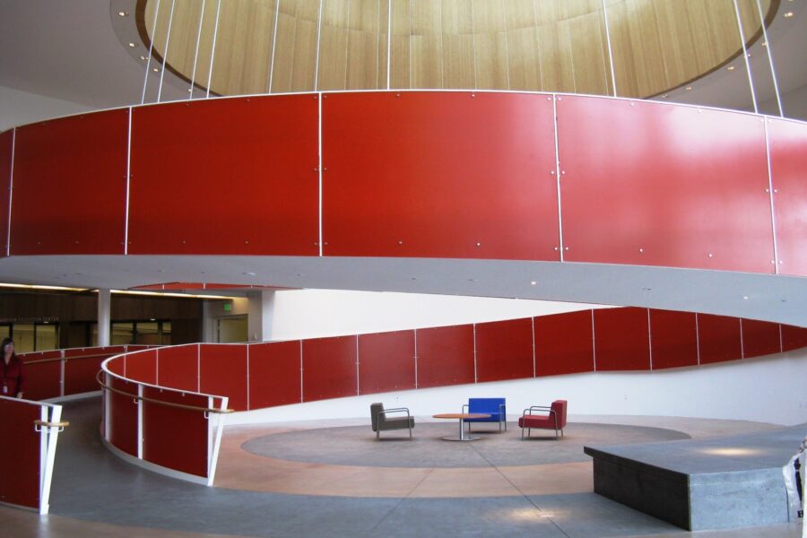 Mynd sem tekin er innandyra í stóru rými. Breiður, rauður spíralrampi hringast upp og liggur á milli tveggja hæða. Undir rampinum eru nokkrir stólar og borð á opinu svæði. Fyrir ofan rampinn er stórt hringlaga ljósop í klædd við, sem hleypir birtu niður í rýmið.