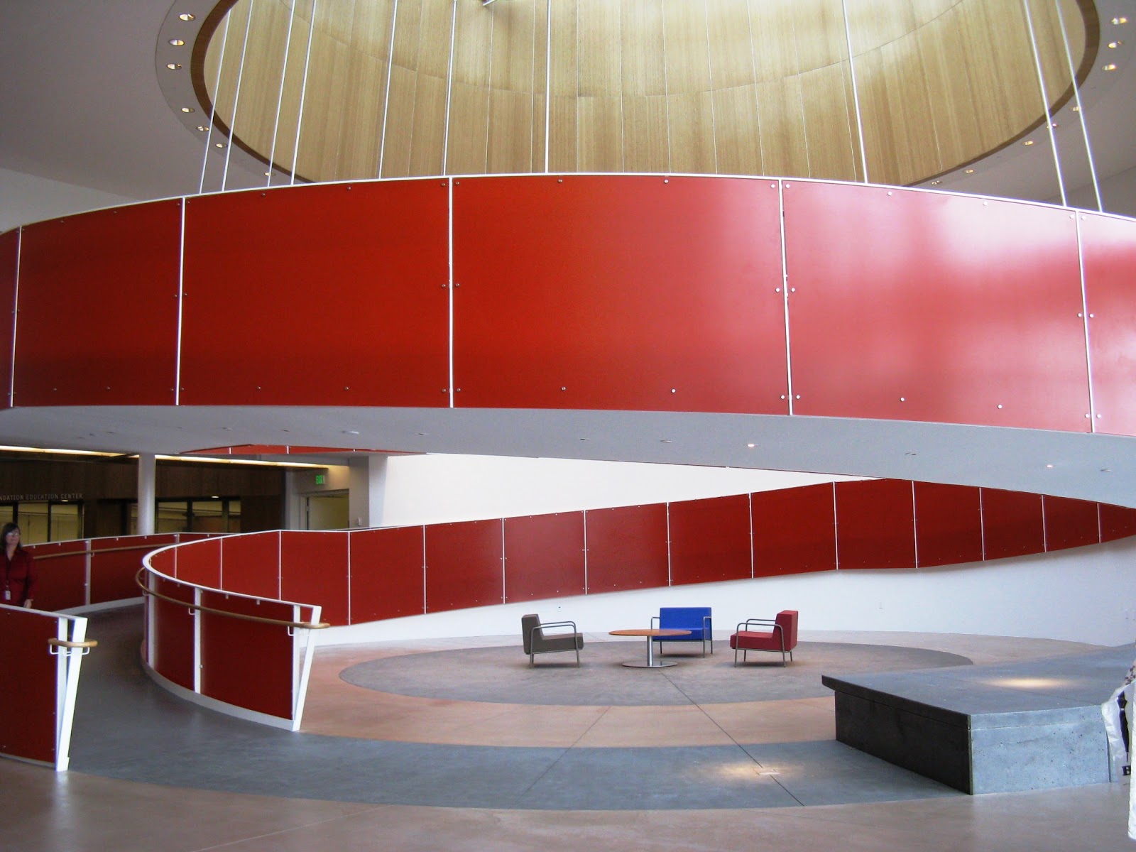 Ed Roberts Campus í Kaliforníu Mynd sem tekin er innandyra í stóru rými. Breiður, rauður spíralrampi hringast upp og liggur á milli tveggja hæða. Undir rampinum eru nokkrir stólar og borð á opinu svæði. Fyrir ofan rampinn er stórt hringlaga ljósop í klædd við, sem hleypir birtu niður í rýmið.