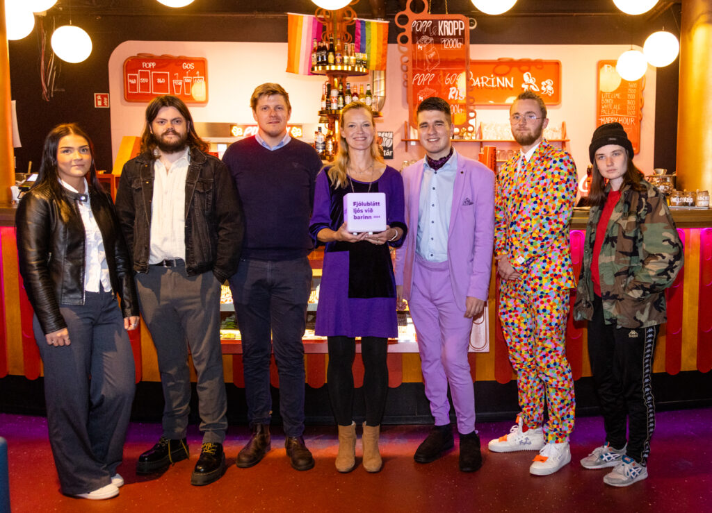 Fjólublátt ljós við barinn Hópmynd af fólki í Bíó Paradís. Þau standa við barinn. Í bakgrunni sést í regnbogafána.