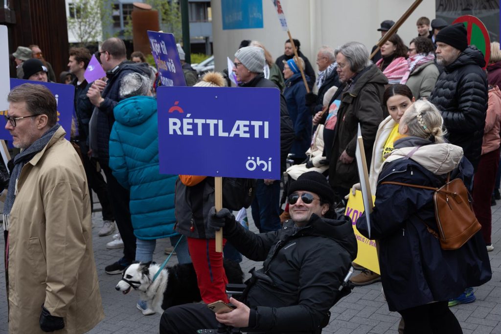 Á myndinni er hópur fólks á Ingólfstorgi á 1. maí 2025. Flest eru í hlýjum vetrarfötum og margir bera skilti. Í forgrunni situr brosandi karlmaður í hjólastól, klæddur dökkum fatnaði og með sólgleraugu. Hann heldur á fjólubláu skilti sem á stendur „RÉTTLÆTI“ ásamt merki ÖBÍ réttindasamtaka. Í kringum hann er fleira fólk. Sumir með kröfuskilti og sumir að tala saman eða fylgjast með. Stemningin á myndinni virðist samheldin og baráttuglöð, þar sem fjölbreyttur hópur fólks tekur þátt.