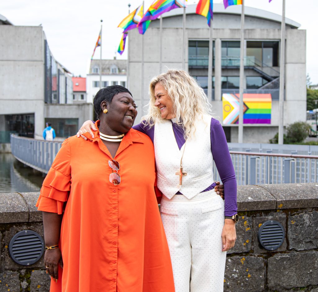 Lady Phyll hjá UK Black Pride og Guðrún Karls Helgudóttir biskup Íslands, fyrir framan ráðhúsið. Þær horfa brosandi á og halda utan um hvror aðra. Ráðhúsið í baksýn og og regnbogafánar blakta.