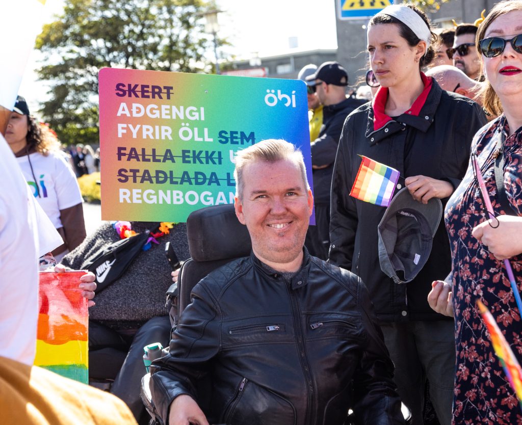 Maður í hjólastól brosir til myndavélarinnar á Gleðigöngu. Hann er í svörtum leðurjakka og í kringum hann er fjölmenni með regnbogafána og skiltum. Bakvið hann sést skilti í regnbogalitum frá ÖBÍ með textanum: „Skert aðgengi fyrir öll sem falla ekki undir staðlaða liti í regnboganum!“ Sólin skín og stemning.
