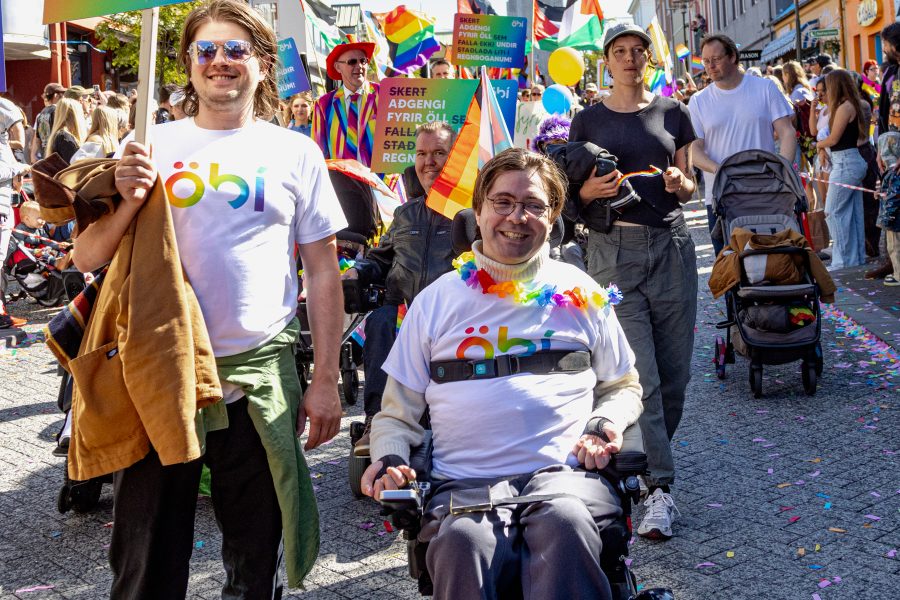 Myndin er tekin í Gleðigöngunni 2025. Í forgrunni eru tveir fulltrúar ÖBÍ réttindasamtaka. Annar þeirra, Rúnar Björn Herrera formaður NPA samtakanna, situr í rafmagnshjólastól, brosandi, og er í bol merktum ÖBÍ. Hann er einnig með regnbogalitað skraut um hálsinn. Við hlið hans stendur maður í samskonar öbÍ bol. Hann heldur á skilti sem á stendur: Skert aðgengi! Fyrir öll sem falla ekki undir staðalbúnað í mannréttindamálum. Í kringum þá eru margir þátttakendur í göngunni, margir með regnbogafána og litrík föt. Göngufólkið gengur niður Skólavörðustíg og Hallgrímskirkja, er í baksýn. Gatan er máluð í regnbogalitum og hátíðarstemning ríkir í mannhafinu.