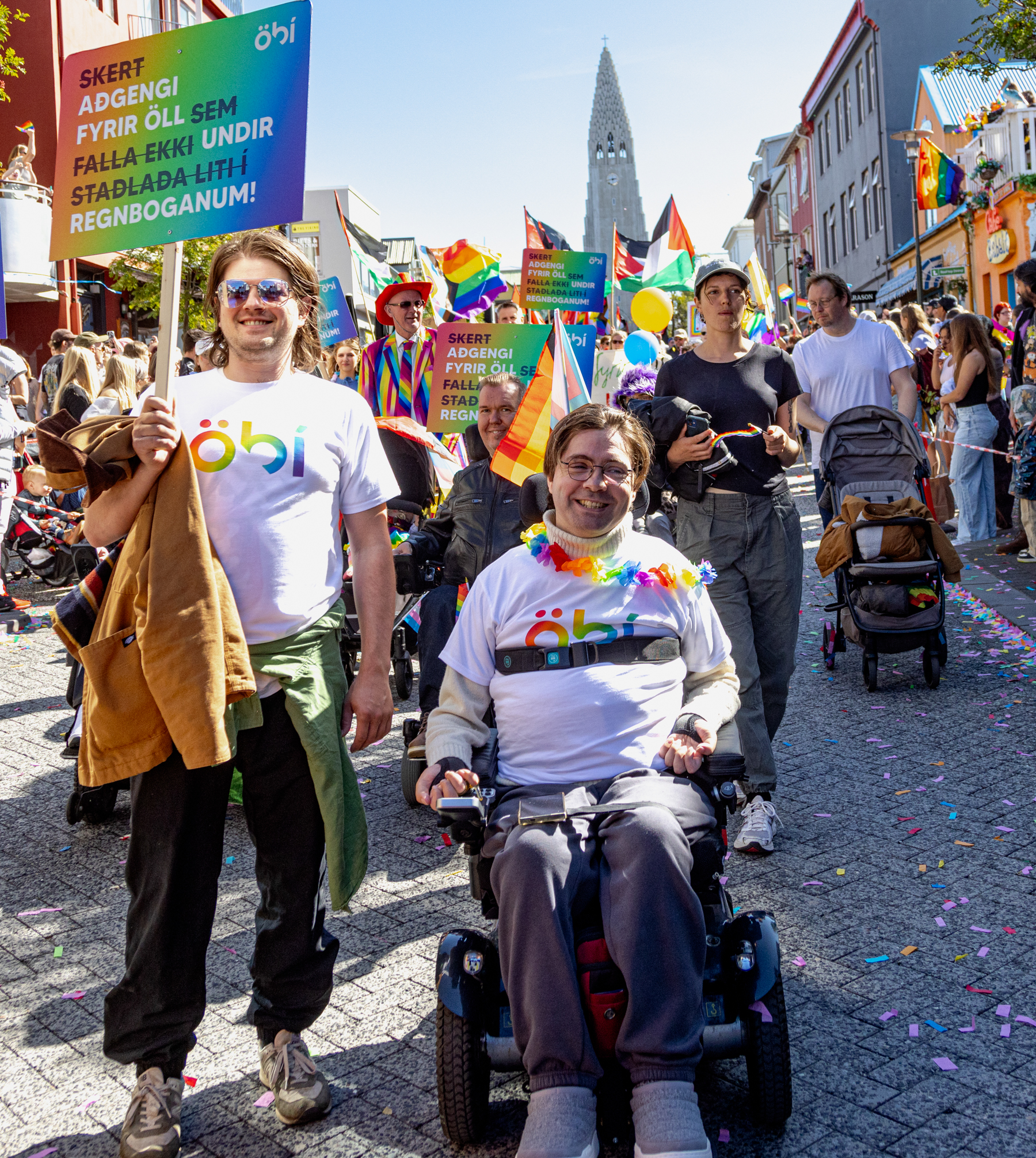 Gleðigangan Myndin er tekin í Gleðigöngunni 2025. Í forgrunni eru tveir fulltrúar ÖBÍ réttindasamtaka. Annar þeirra, Rúnar Björn Herrera formaður NPA samtakanna, situr í rafmagnshjólastól, brosandi, og er í bol merktum ÖBÍ. Hann er einnig með regnbogalitað skraut um hálsinn. Við hlið hans stendur maður í samskonar öbÍ bol. Hann heldur á skilti sem á stendur: Skert aðgengi! Fyrir öll sem falla ekki undir staðalbúnað í mannréttindamálum. Í kringum þá eru margir þátttakendur í göngunni, margir með regnbogafána og litrík föt. Göngufólkið gengur niður Skólavörðustíg og Hallgrímskirkja, er í baksýn. Gatan er máluð í regnbogalitum og hátíðarstemning ríkir í mannhafinu.