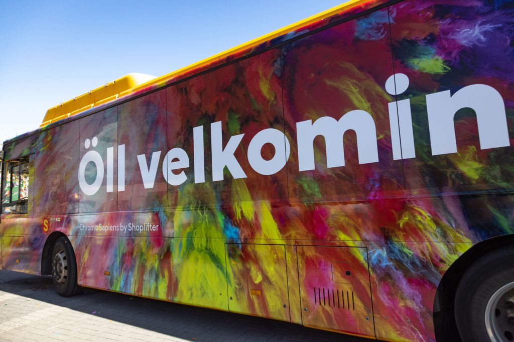 Strætisvagn skreyttur litríkum mynstrum í regnbogalitum með stórum hvítum stöfum á hliðinni sem mynda orðin „Öll velkomin“. Undir stendur „Chromo Sapiens by Shoplifter“. Himinninn er heiðblár og sólin skín.