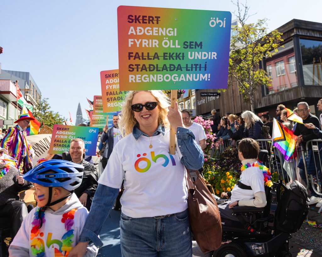 Hópur fólks úr ÖBÍ réttindasamtökum tekur þátt í Gleðigöngunni fyrir framan Hallgrímskirkju. Þau bera skilti með textanum „Skert aðgengi fyrir öll sem falla ekki undir staðlaða liti regnbogans!“ Formaður ÖBÍ, Alma Ýr Ingólfsdóttir fremst í broddi fylkningar ásamt syni sínum.