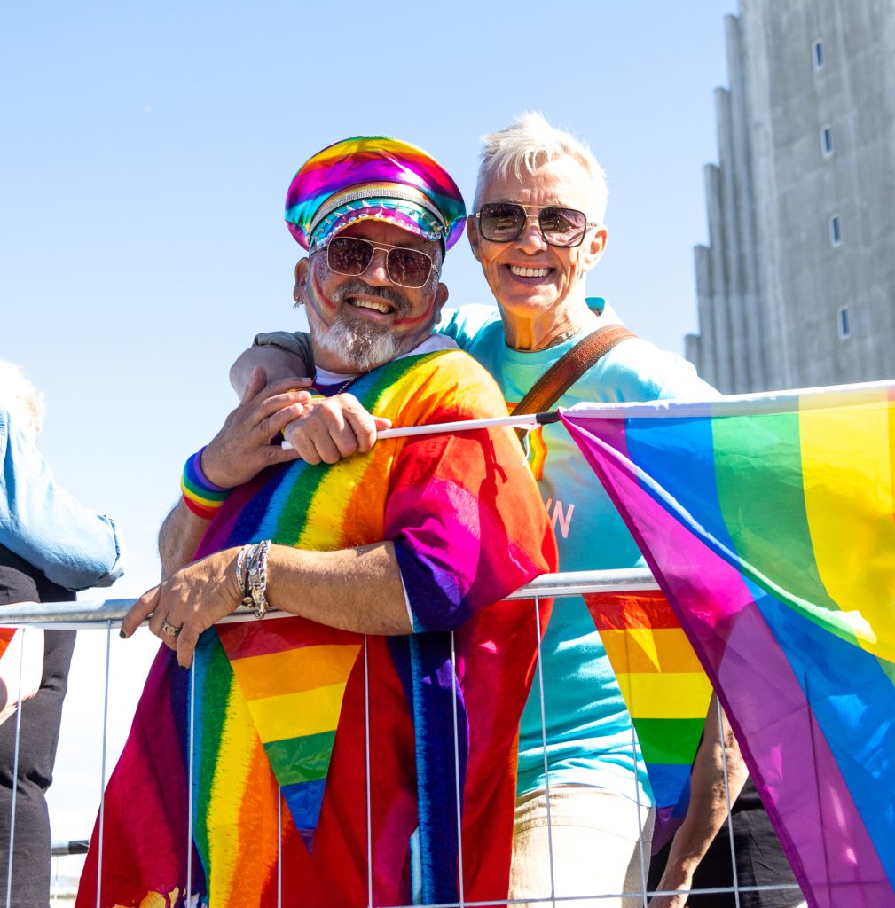Tveir menn standa hlið við hlið og brosa breitt í sólskini á Gleðigöngu. Annar er í litríkum regnbogabúningi og samsvarandi húfu, hinn í ljósblárri peysu með sólgleraugu. Þeir halda á regnbogafána og faðmast yfir grindverki. Í bakgrunni sést Hallgrímskirkja.
