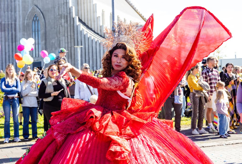Dragdrottning í glæsilegum rauðum kjól með stórum vængjum og skreyttu kórónu dansar í Gleðigöngunni fyrir framan Hallgrímskirkju. Í bakgrunni fylgist hópur áhorfenda með, sumir halda á litríkum blöðrum og regnbogafánum.