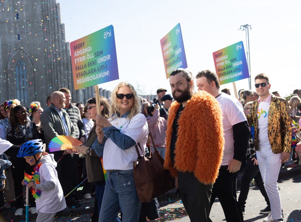 Hópur fólks úr ÖBÍ réttindasamtökum tekur þátt í Gleðigöngunni fyrir framan Hallgrímskirkju. Þau bera skilti með textanum „Skert aðgengi fyrir öll sem falla ekki undir staðlaða liti regnbogans!“ Fremst í broddi fylkningar er formaður ÖBÍ, Alma Ýr og Þórgnýr Einar upplýsingafulltrúi ÖBÍ.