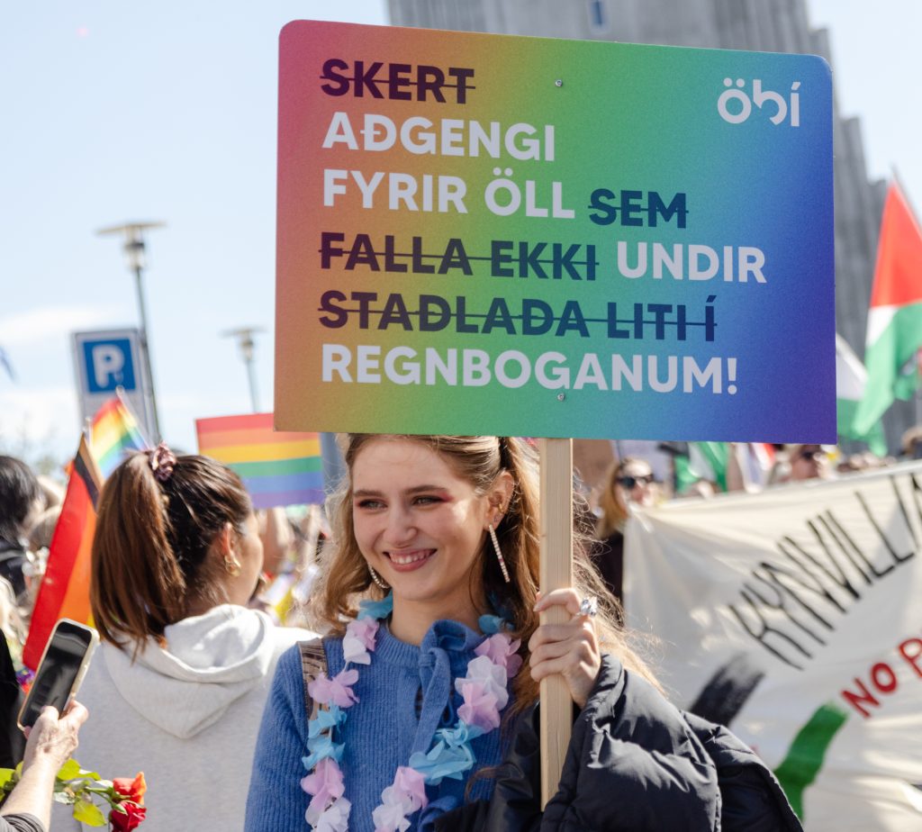 Ung kona brosir og heldur á skilti í Gleðigöngunni með áletruninni: „Skert aðgengi fyrir öll sem falla ekki undir staðlaða liti í regnboganum!“ Skiltið er í regnbogalitum með merki ÖBÍ. Í bakgrunni sjást þátttakendur með fána og borða.