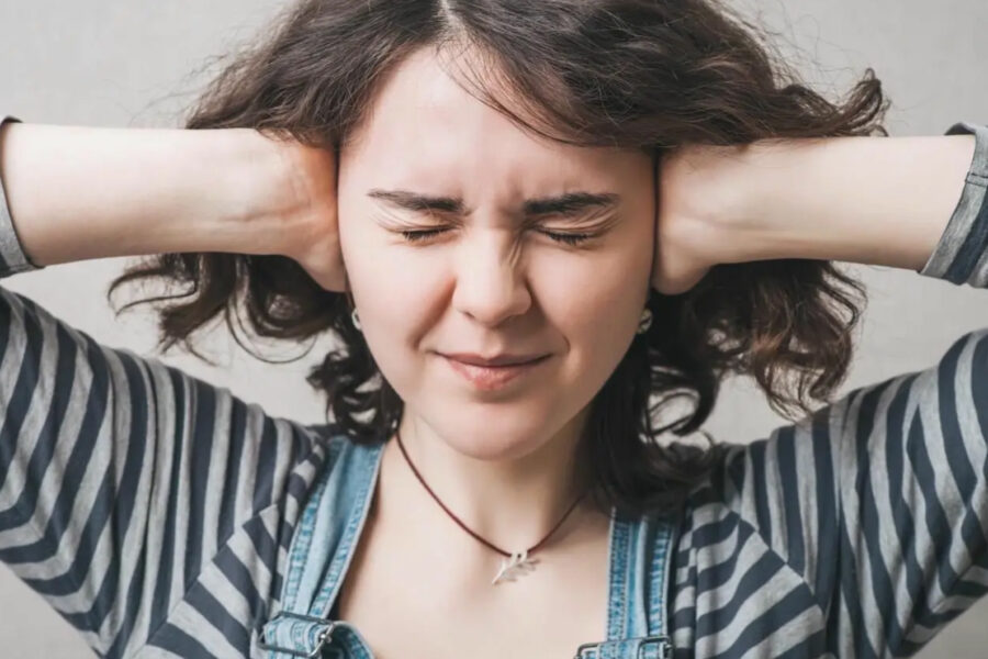 Nærmynd af konusem heldur með báðum höndum utan um eyrun, með lokuð augun. Peysan sem hún klæðist er röndótt sem er tilvísun í gangbrautir og umferðarhávaða.