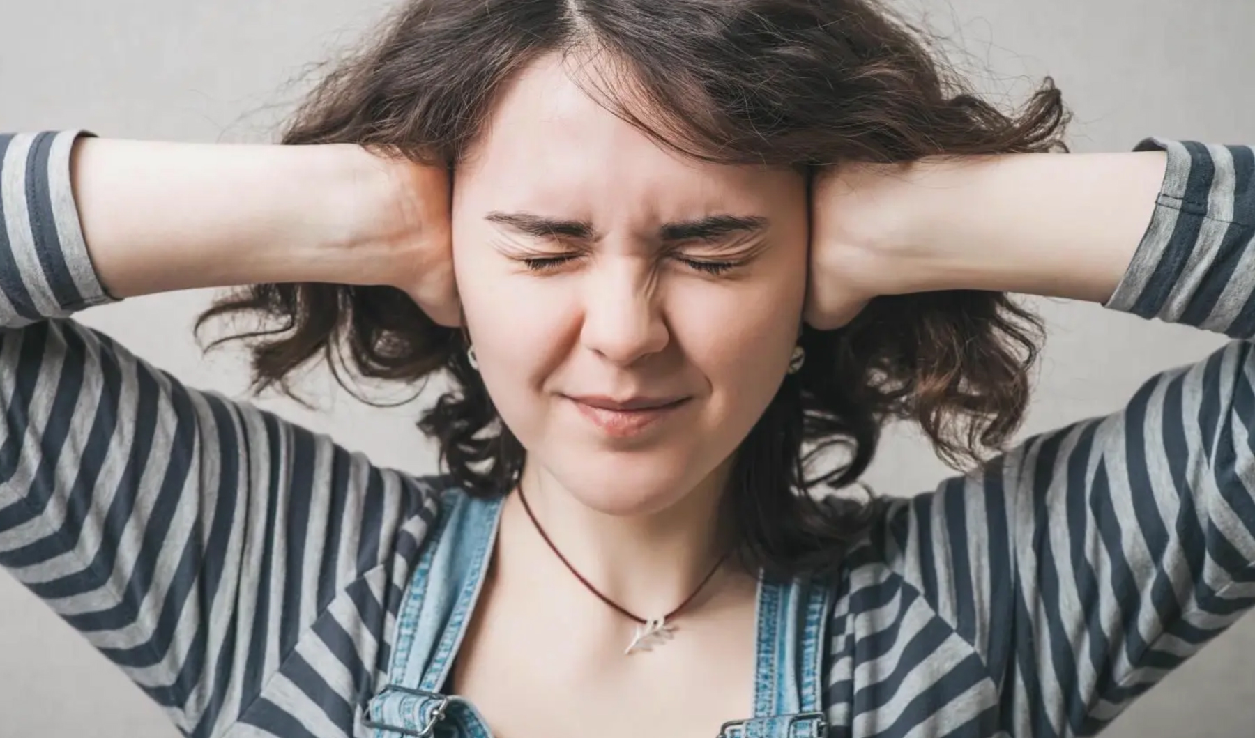 Nærmynd af konusem heldur með báðum höndum utan um eyrun, með lokuð augun. Peysan sem hún klæðist er röndótt sem er tilvísun í gangbrautir og umferðarhávaða.