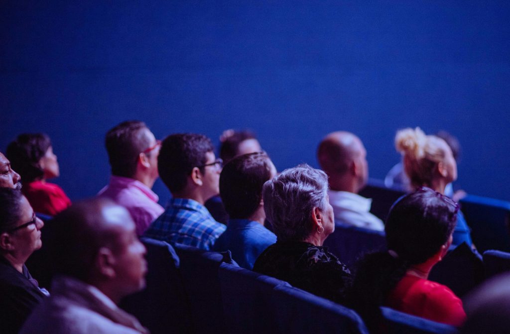 „Hópur fólks situr í röðum í sal og horfir fram á við, líklega á fyrirlestur. Myndin er tekin aftan frá og sýnir fólk af ólíkum aldri, þjóðerni og kyni í . áhorfendastæðum.“