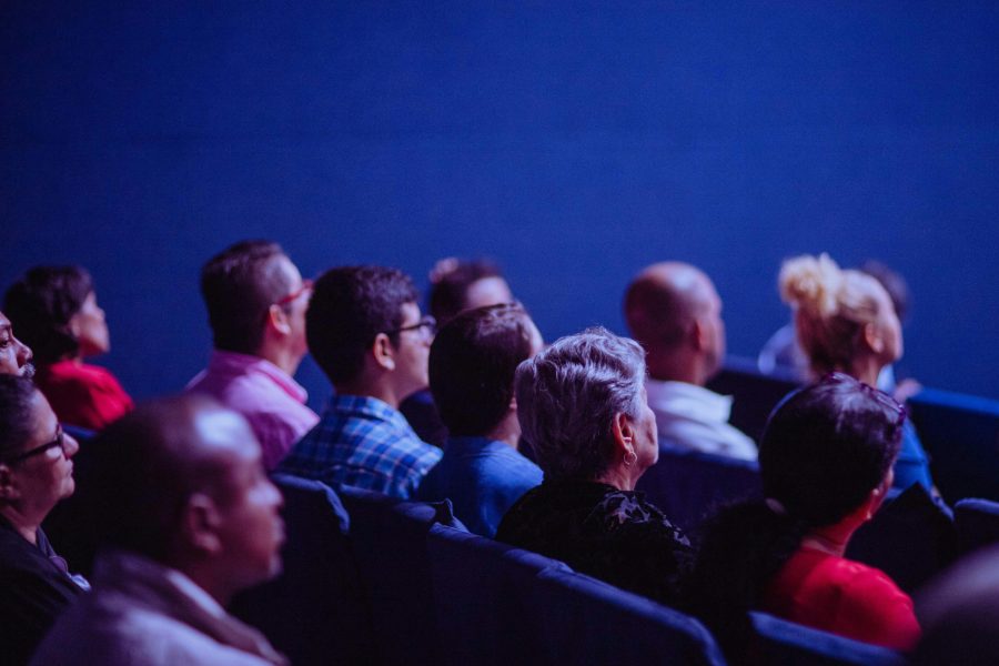 „Hópur fólks situr í röðum í sal og horfir fram á við, líklega á fyrirlestur. Myndin er tekin aftan frá og sýnir fólk af ólíkum aldri, þjóðerni og kyni í . áhorfendastæðum.“
