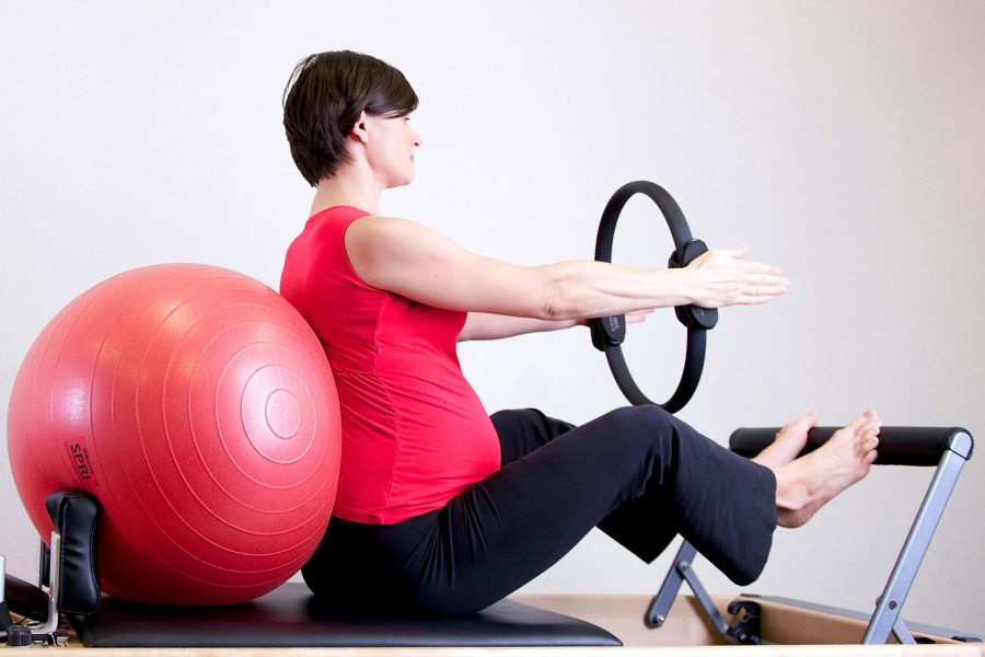 „Barnshafandi kona situr á Pilates-reformer tæki með æfingabolta við bakið og heldur á Pilates-hring.“