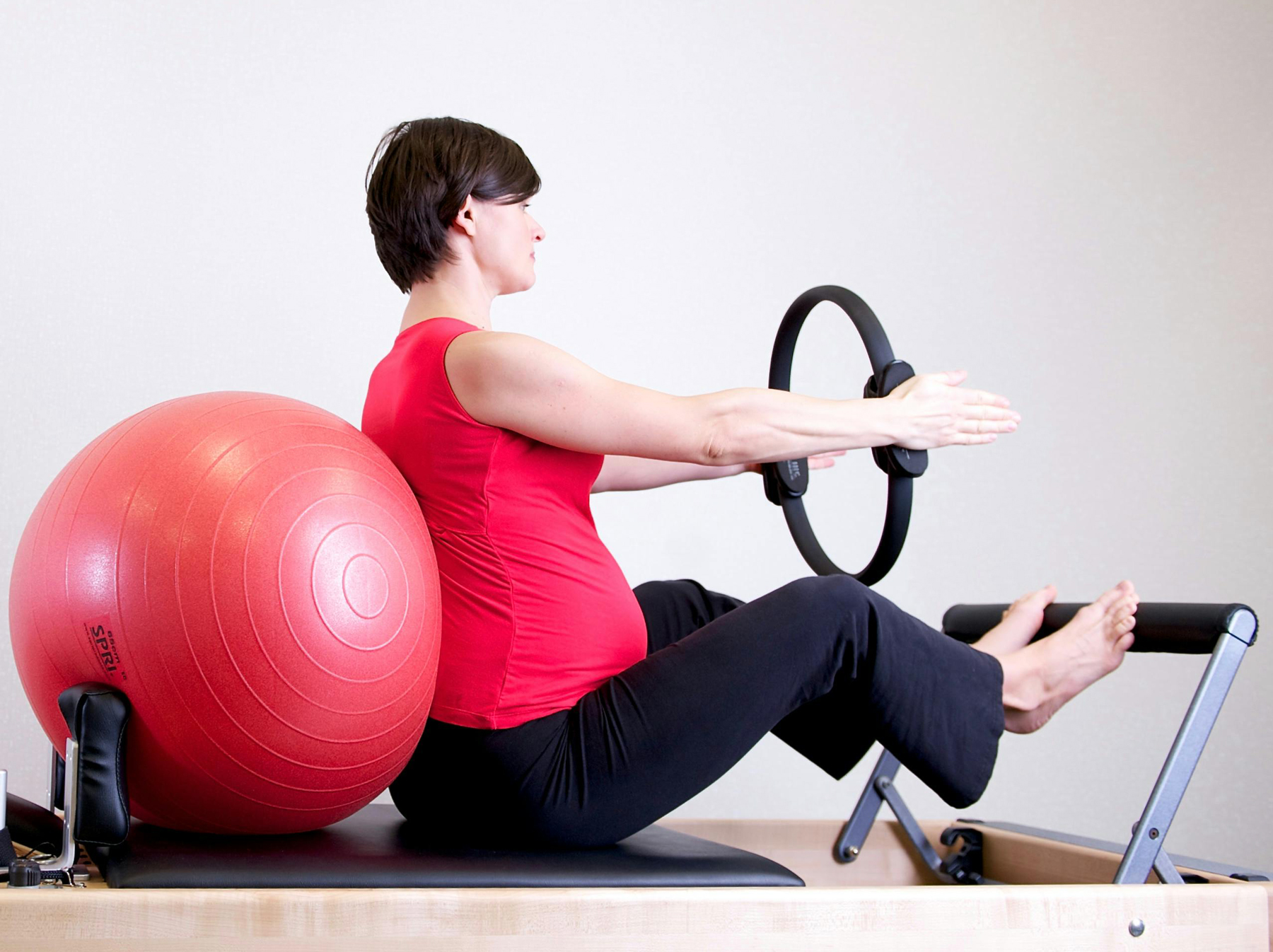 „Barnshafandi kona situr á Pilates-reformer tæki með æfingabolta við bakið og heldur á Pilates-hring.“