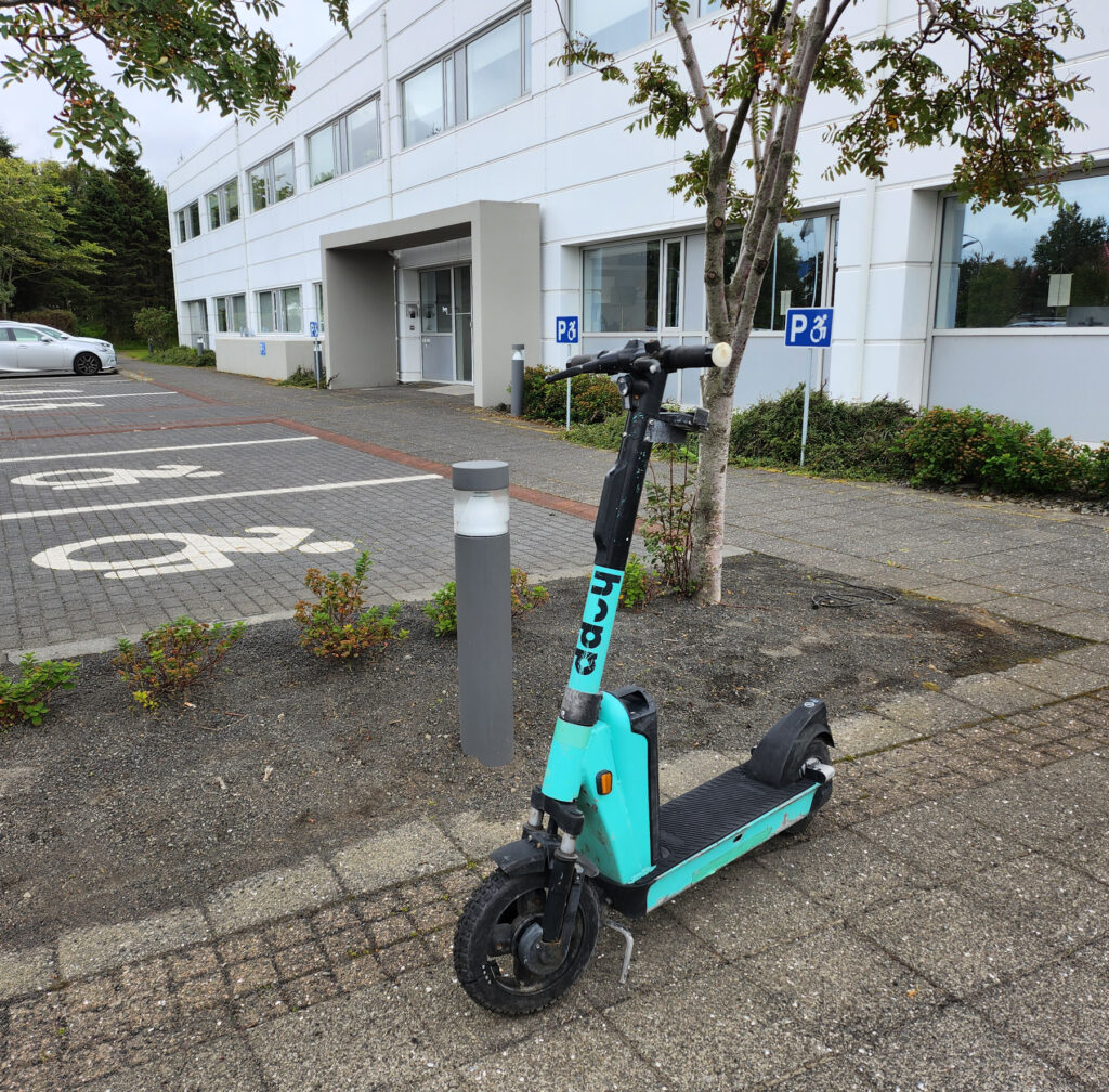 Hopp rafskúta stendur á gangstétt við hlið bílastæða merktum fyrir hreyfihamlaða og hindrar aðgengi að leiðinni frá stæðunum að inngangi hússins.