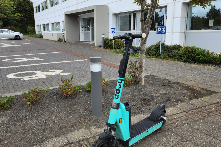 Hopp rafskúta stendur á gangstétt við hlið bílastæða merktum fyrir hreyfihamlaða og hindrar aðgengi að leiðinni frá stæðunum að inngangi hússins.