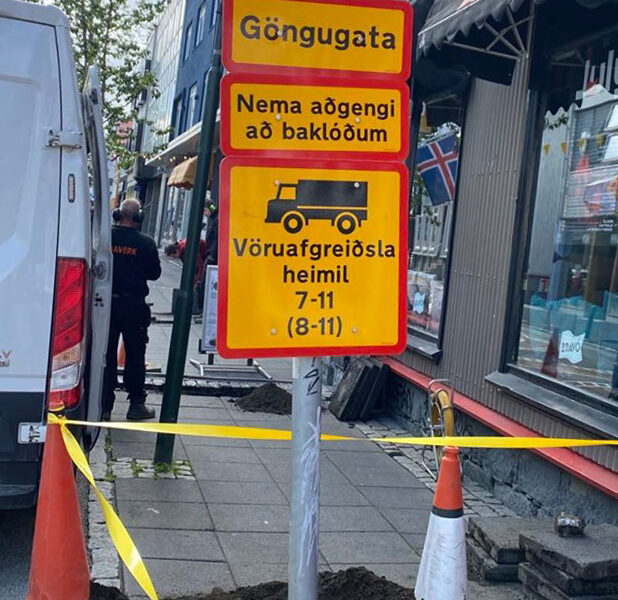 Ljósmyndin sýnir: „Gult og rautt umferðarmerki á gangstétt með textanum: Göngugata. Nema aðgengi að baklóðum. Vöruafgreiðsla heimil 7–11 (8–11). Undir merkinu má sjá vinnusvæði afmarkað með keilum og gulum borðum.“