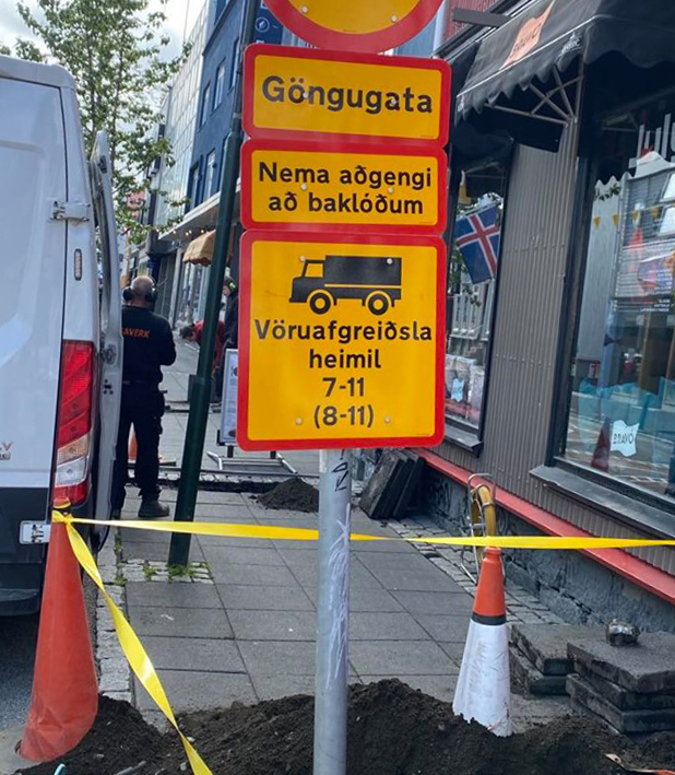 Ljósmyndin sýnir: „Gult og rautt umferðarmerki á gangstétt með textanum: Göngugata. Nema aðgengi að baklóðum. Vöruafgreiðsla heimil 7–11 (8–11). Undir merkinu má sjá vinnusvæði afmarkað með keilum og gulum borðum.“