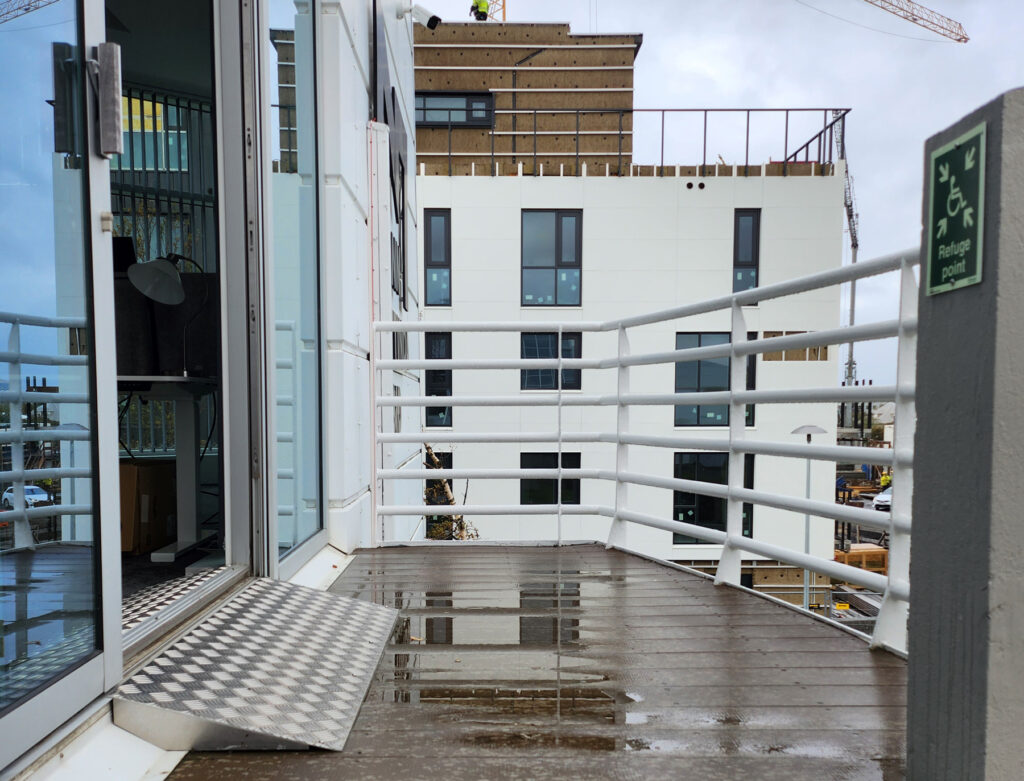 Á myndinni sést neyðarútgangaopnun út á svalir í skrifstofubyggingu. Við útganginn er settur hallandi álkanti sem myndar ramp yfir þröskuld til að auðvelda fólki í hjólastól eða með aðra hreyfihömlun að komast á flóttasvæði á annari hæð.