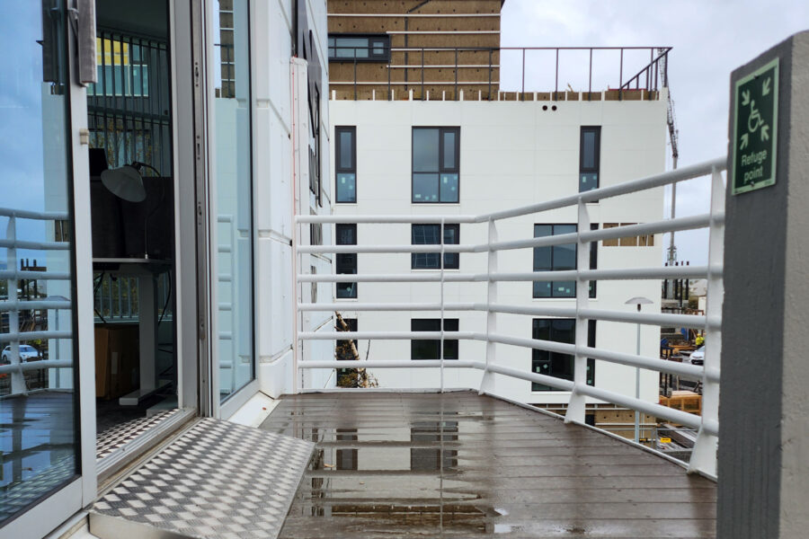 Á myndinni sést neyðarútgangaopnun út á svalir í skrifstofubyggingu. Við útganginn er settur hallandi álkanti sem myndar ramp yfir þröskuld til að auðvelda fólki í hjólastól eða með aðra hreyfihömlun að komast á flóttasvæði á annari hæð.