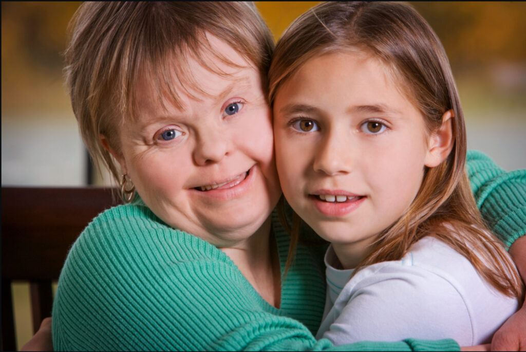 „Móðir með Downs heilkenni og dóttir hennar sitja þétt saman og faðmast. Þær horfa báðar beint í myndavélina.