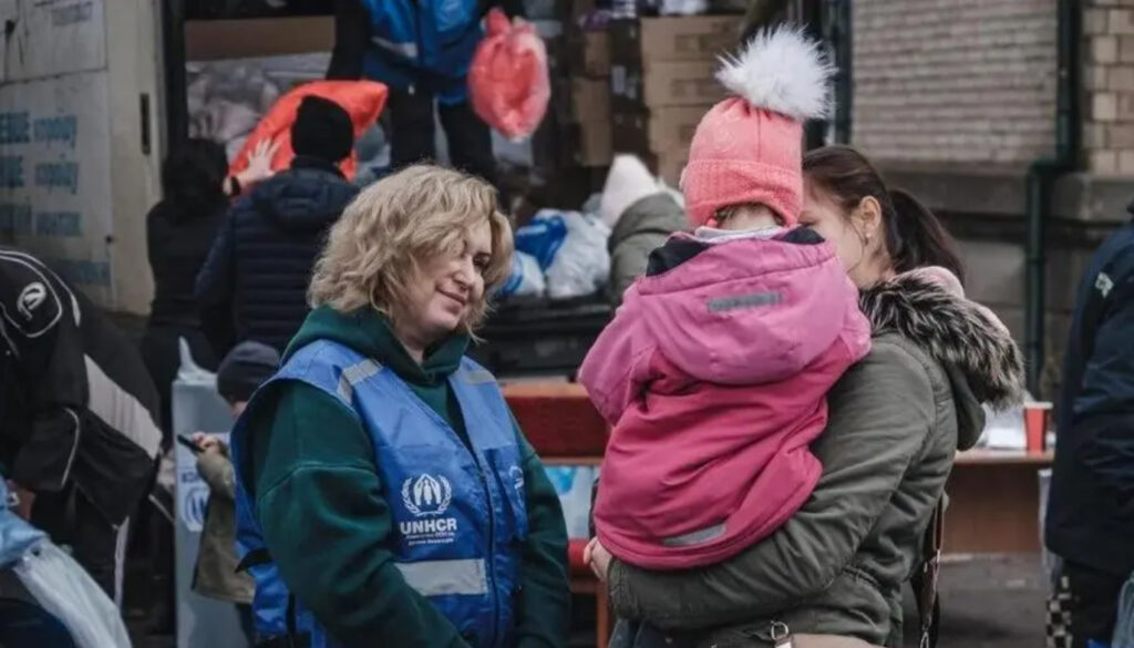 Kona í vesti merkt UNHCR stendur úti við hjálpardreifingu og talar hlýlega við móður sem heldur á barni í vetrarfötum. Í bakgrunni sjást fleiri sjálfboðaliðar og sendibíll með kassa og poka af hjálpargögnum.