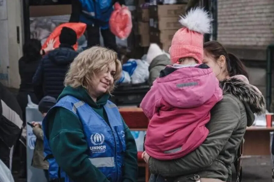 Kona í vesti merkt UNHCR stendur úti við hjálpardreifingu og talar hlýlega við móður sem heldur á barni í vetrarfötum. Í bakgrunni sjást fleiri sjálfboðaliðar og sendibíll með kassa og poka af hjálpargögnum.