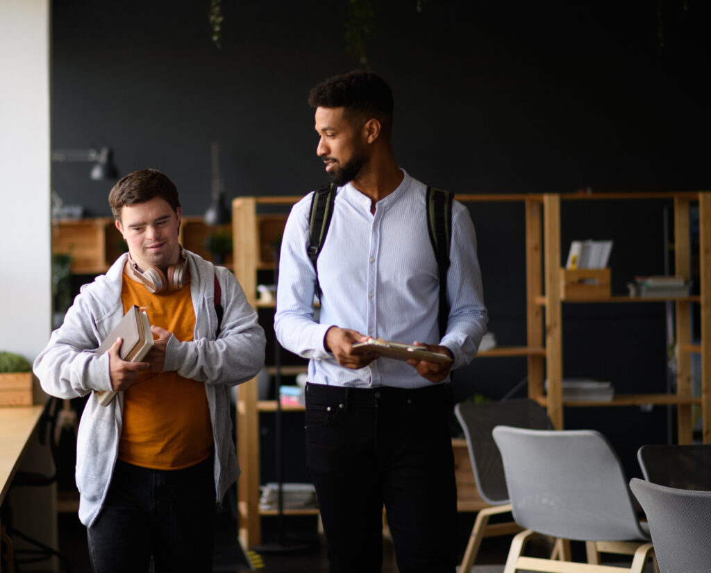 Kennari og nemandi (með Downs heilkenni) , ganga saman innandyra og halda á bókum og spjaldtölvu.