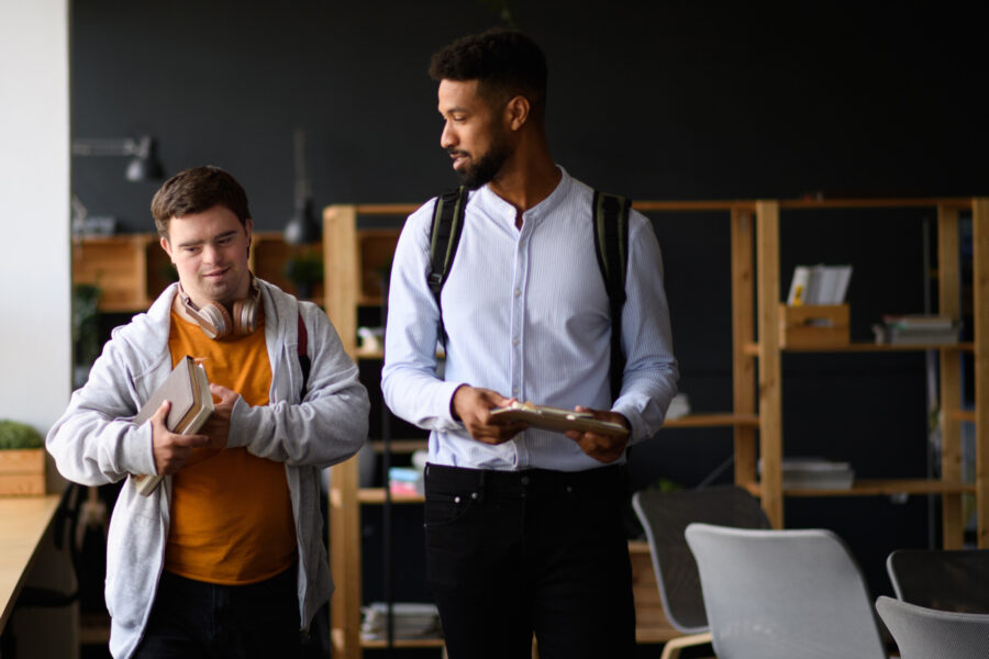 Kennari og nemandi (með Downs heilkenni) , ganga saman innandyra og halda á bókum og spjaldtölvu.