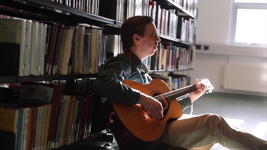 Maður situr við bókaskáp með gítar