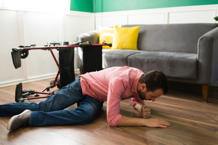 Maður liggur á gólfi inni í stofu, með hendurnar yfir höfði í verndarstöðu eins og mælt er með í jarðskjálfta („drop, cover, hold“). Við hlið hans er hjólastóll sem hefur oltið um koll.