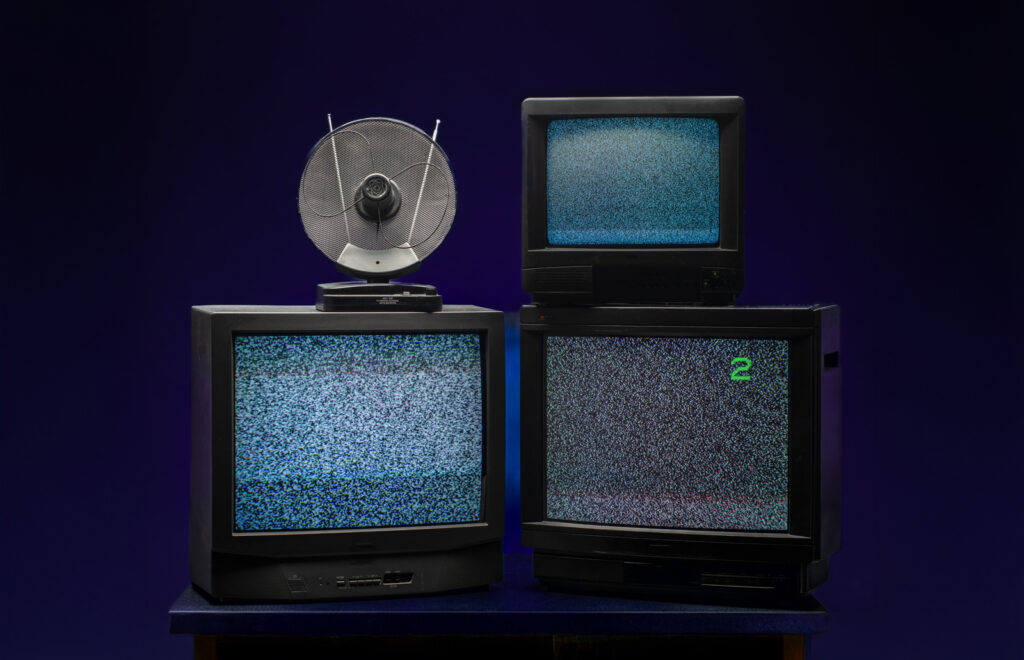 Þrír gamlir kassalaga sjónvarpsskjáir standa staflaðir á borði með truflunum á skjánum; ofan á einum er lítið loftnet.