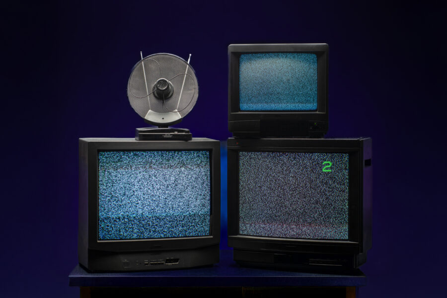 Þrír gamlir kassalaga sjónvarpsskjáir standa staflaðir á borði með truflunum á skjánum; ofan á einum er lítið loftnet.