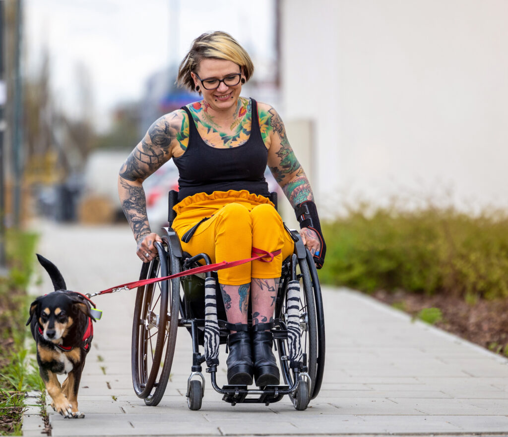 Kona í hjólastól rúllar eftir gangstétt, og leiðir lítinn hund í taum. Hún er glaðbeitt og handleggirnir er litskrúðugir af tattúum.