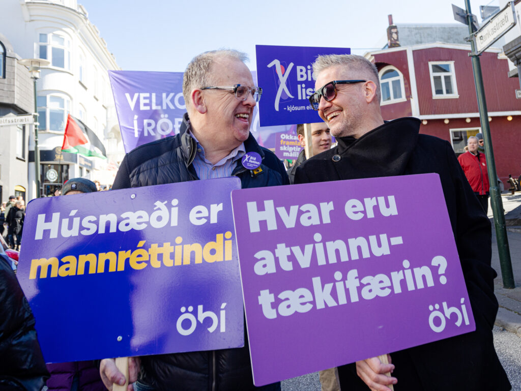 Tvær manneskjur standa hlið við hlið í 1. maí réttindagöngu og brosa hvor til annarrar. Þær halda hvor skiltum merktum ÖBÍ. 1.sta skiltið með textanum: „Húsnæði er mannréttindi“ og á hinu er textinn: Hvar eru atvinnutækifærin.