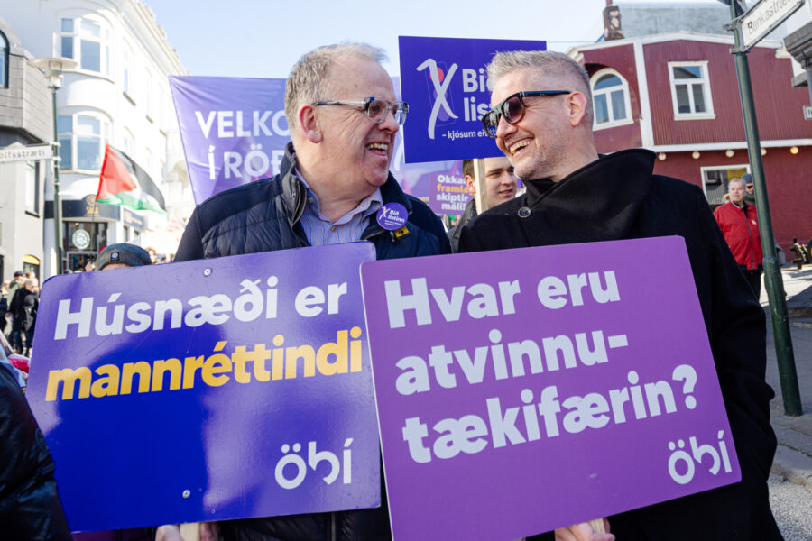 Tvær manneskjur standa hlið við hlið í 1. maí réttindagöngu og brosa hvor til annarrar. Þær halda hvor skiltum merktum ÖBÍ. 1.sta skiltið með textanum: „Húsnæði er mannréttindi“ og á hinu er textinn: Hvar eru atvinnutækifærin.